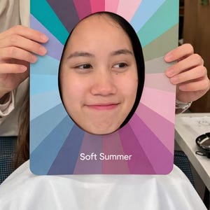 May include: A color analysis tool with a cutout for a face, featuring a spectrum of soft summer colors. The tool is held up in front of a person, with the text "Soft Summer" displayed at the bottom.
