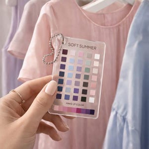 Pode incluir: Uma paleta de cores de plástico transparente com a etiqueta "SOFT SUMMER" com uma variedade de amostras de cores pastel. A paleta é segurada em frente a um varal de roupas em tons de rosa, lavanda e azul. As palavras "MAKE UP COLOR" estão impressas na paleta.