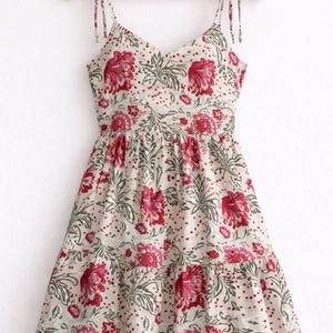 May include: A cream-colored sundress with a floral pattern of red flowers and green leaves. The dress has a V-neck, tie straps, and a tiered skirt. It is hanging on a wooden hanger.
