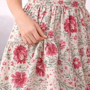 May include: A floral print dress featuring large pink flowers and smaller red flowers on a cream background. The dress has a tiered skirt and a fitted bodice. The fabric has a delicate, vintage look, perfect for a summer day.