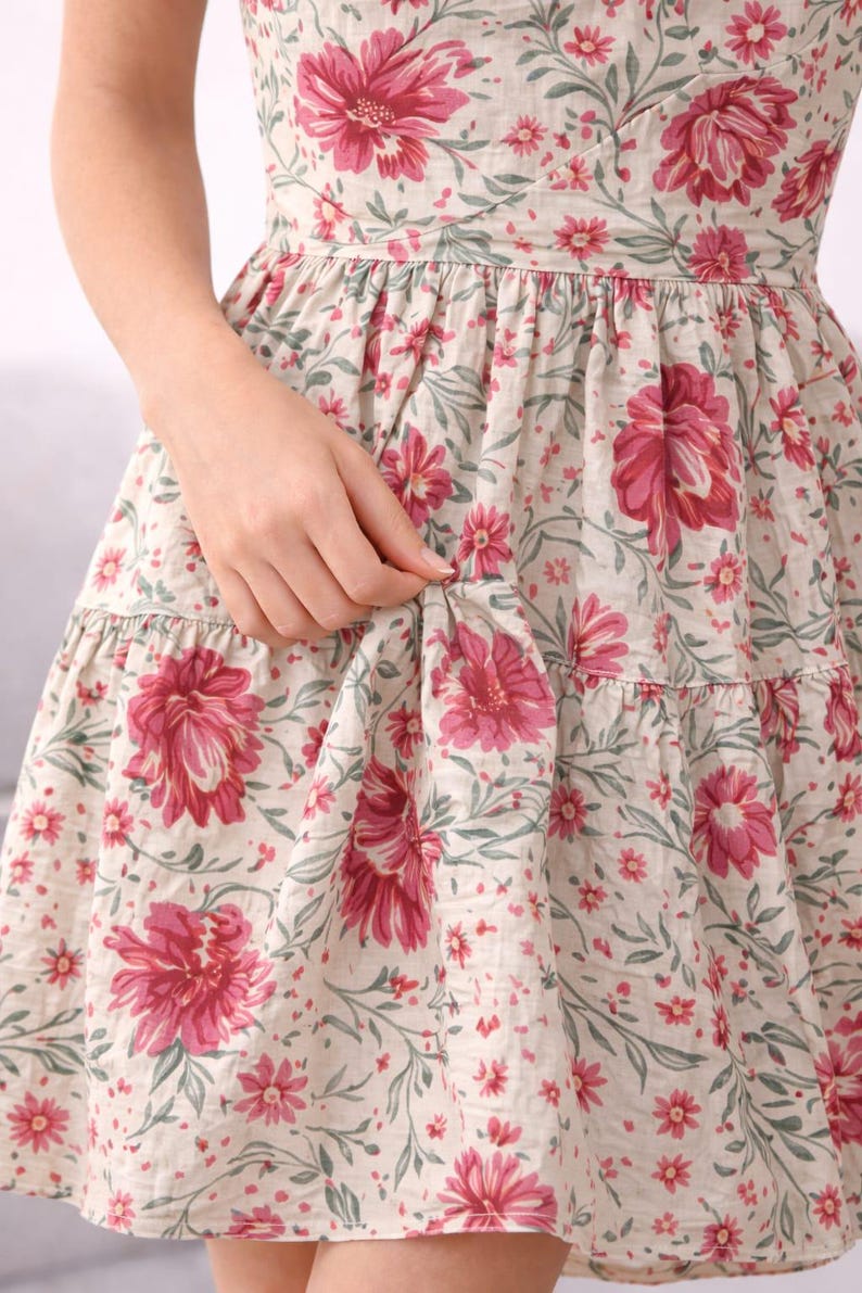May include: A floral print dress featuring large pink flowers and smaller red flowers on a cream background. The dress has a tiered skirt and a fitted bodice. The fabric has a delicate, vintage look, perfect for a summer day.