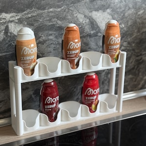 Könnte beinhalten: Ein weißer Kunststoff-Organizer mit fünf Flaschen More Zerup-Sirup. Die Flaschen sind in verschiedenen Farben gehalten, darunter Braun und Rot, mit weißen Deckeln. Die Sirups sind für Kaffee und Tee. Der Organizer steht auf einer Arbeitsplatte.