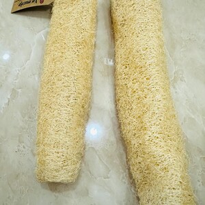 May include: Two natural loofah sponges, cylindrical in shape, are displayed on a marble surface. The sponges have a light beige color and a fibrous texture. One sponge has a tag attached with a string.