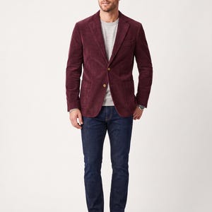 Pode incluir: Um blazer de veludo cotelê bordô escuro com dois botões frontais e bolsos chapados, usado sobre uma camiseta cinza claro de gola redonda, combinado com jeans azul escuro e botas chukka de couro marrom. O blazer possui lapelas entalhadas.