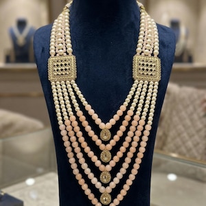 May include: A multi-strand necklace displayed on a dark blue mannequin. The necklace features strands of white and peach-colored beads, with gold-colored square accents and teardrop pendants. The necklace is a statement piece.