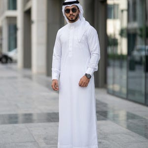 Może przedstawiać: Mężczyzna w tradycyjnym białym thobe, długiej, luźnej szacie, oraz czarno-białym nakryciu głowy keffiyeh. Ma na sobie okulary przeciwsłoneczne i zegarek na rękę. Tło przedstawia nowoczesny budynek.