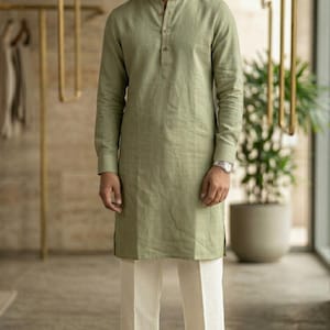 May include: A man wearing a long-sleeved, olive green tunic shirt with a mandarin collar and button closure, paired with off-white linen pants. He is also wearing a silver watch and brown leather sandals. The outfit is a casual, yet stylish ensemble.