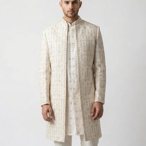 May include: Cream-colored, long embroidered coat with a mandarin collar, worn over a matching tunic and pants. The coat features a detailed geometric pattern in a lighter shade. The outfit is a formal ensemble.