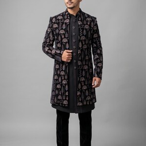 Könnte beinhalten: Ein schwarzer Samt-Langmantel mit floralem Muster, getragen über einem schwarzen Hemd und einer Hose. Der Mantel hat einen Stehkragen und einen Knopfverschluss. Das Outfit wird durch schwarze Anzugschuhe ergänzt.
