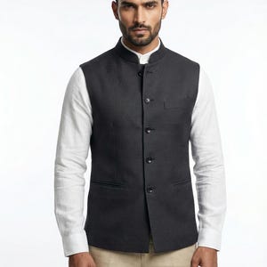 Puede incluir: Un chaleco gris oscuro con botones y cuello mandarín. El chaleco tiene un corte a medida y presenta un bolsillo en el pecho y dos bolsillos delanteros. El chaleco se usa sobre una camisa blanca y se combina con pantalones color beige.