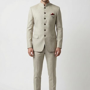 May include: A light beige suit with a mandarin collar, featuring a matching jacket and trousers. The jacket has a pocket square and a decorative pin. The suit is paired with brown leather shoes, creating a formal ensemble.