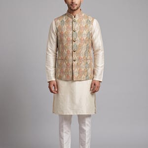 Op de afbeelding: Een crèmekleurige kurta-pyjamaset met een patroonvest. Het vest heeft een herhalend ontwerp in tinten beige, groen en oranje. De outfit bevat een shirt met lange mouwen en een witte broek. Bruine leren schoenen maken de set compleet.