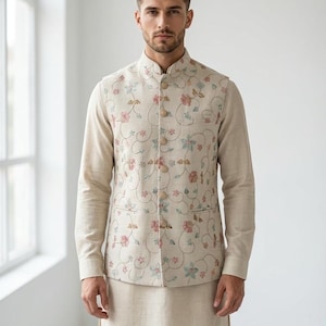May include: Beige floral embroidered vest with matching long-sleeve shirt and pants. The vest features a mandarin collar and button closure. The floral embroidery includes pink, blue, and green accents. The outfit is a neutral color.