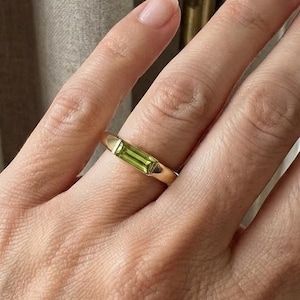 Puede incluir: Un anillo de oro con una piedra preciosa rectangular de color verde claro en talla baguette, engastada horizontalmente en el centro de la banda. El anillo de metal tiene un acabado liso y pulido con un diseño sencillo que destaca la piedra.