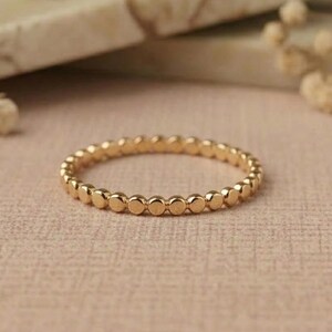 May include: A thin gold ring featuring a continuous band of small, uniform, spherical beads arranged in a circular pattern. The metal has a polished, reflective finish that catches the light, creating a simple and consistent design.