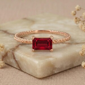 Puede incluir: Un anillo de oro rosa con una piedra preciosa roja de corte esmeralda rectangular montada en cuatro garras. La banda del anillo presenta una textura trenzada similar a una cuerda, lo que aporta un detalle distintivo a la superficie metálica.