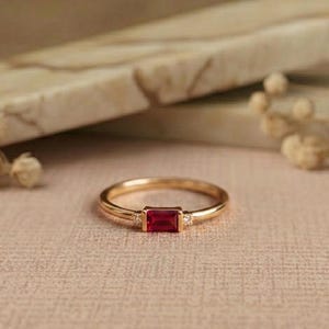 May include: A gold ring featuring a central rectangular baguette-cut red gemstone flanked by two small clear round stones on either side. The band is thin and polished, showcasing a clean design with a vibrant red center stone and clear accents.