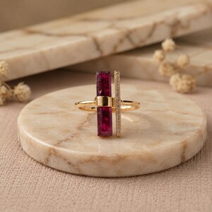 May include: A gold ring featuring a vertical rectangular deep magenta gemstone and a parallel thin gold bar encrusted with small clear stones. The ring band is thin and polished gold, creating a geometric design with contrasting textures.
