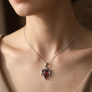 May include: A silver necklace featuring a kite-shaped red gemstone pendant. The pendant is accented with small black stones and two silver crescent-shaped elements on the sides, suspended from a delicate silver chain that rests on the neck.