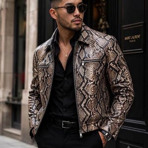 Peut inclure: Une veste en cuir à motif peau de serpent marron et noir avec un col pointu, des manches longues et des fermetures éclair en métal argenté sur les poches avant et la fermeture centrale. La veste est portée sur une chemise noire.