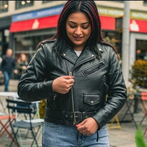 May include: Black leather motorcycle jacket with a belted waist, zippered pockets, and a classic asymmetrical zipper. The jacket is worn with blue jeans. The background shows a street scene with a red awning.