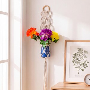 May include: A wall-hanging macrame plant holder featuring a circular wooden ring at the top, intricate off-white knotted cord patterns, and a blue glass vase containing bright orange, purple, and yellow artificial flowers with green stems.