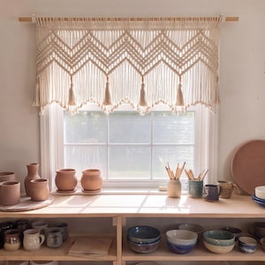May include: An off-white macrame window valance featuring a chevron pattern, knotted fringe, and hanging tassels suspended from a wooden rod. The valance is positioned above a window sill filled with various ceramic pots, bowls, and mugs.