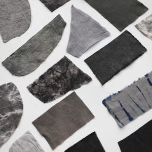 May include: An assortment of irregularly shaped fabric swatches in various shades of gray and black, arranged on a white surface. The textures vary from smooth to heavily textured, with some pieces showing tie-dye patterns and blue accents.