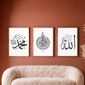 May include: Three framed art prints with black calligraphy on white backgrounds hang above a cream-colored sofa. The prints are in wooden frames and feature Arabic script. The wall is painted a warm, reddish-brown color. A woven basket with tall, dried plants sits to the right.