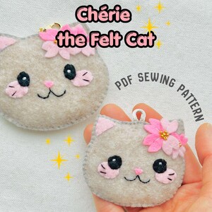 May include: Two felt cat ornaments with pink details and flower decorations. The text "Chérie the Felt Cat" is above the cats, with "PDF Sewing Pattern" below. One cat is held in a hand.