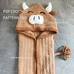Op de afbeelding: Een gehaakte handdoek met capuchon in de vorm van een koe. De handdoek is voornamelijk bruin met bruine randen, hoorns en staart. De capuchon heeft een lachend koeiengezicht met witte hoorns en een witte snuit. De tekst "PDF CROCHET PATTERN ENG" is zichtbaar.