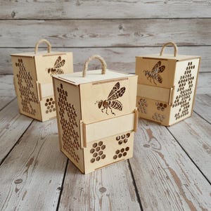 Op de afbeelding: Drie lichtbruine houten dozen met uitgesneden bijen en honingraten. Elke doos heeft een touwen handvat en een lasergesneden bijenontwerp. De dozen staan op een houten oppervlak.