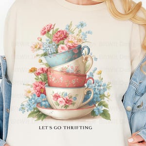 Può includere: Felpa color crema con un design di tazze da tè vintage impilate in colori pastello, decorate con composizioni floreali. Il design include fiori rosa, blu e gialli. Il testo "LET'S GO THRIFTING" è stampato in basso.