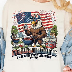 May include: A cream-coloured t-shirt with a patriotic design. It features an eagle wearing sunglasses and a USA hat, grilling and holding a beer. The design includes an American flag, fireworks, and the text "1776 Problems But Freedom Ain't One."