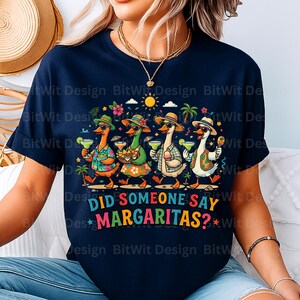 May include: A navy blue t-shirt featuring a graphic of four ducks wearing hats and sunglasses, holding margaritas and snacks. The text reads DID SOMEONE SAY MARGARITAS? in colorful letters. The design includes palm trees, a sun, and tropical flowers.