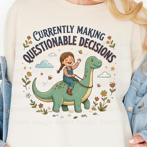 Op de afbeelding: Een crèmekleurig sweatshirt met een afbeelding van een kind met gevlochten haar op een groene dinosaurus. De tekst erboven luidt CURRENTLY MAKING QUESTIONABLE DECISIONS. Het ontwerp bevat oranje bloemen, kleine wolken en vlinders.