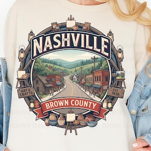 May include: A cream-colored sweatshirt featuring a circular graphic of a town scene with the text Nashville and Brown County. The design includes illustrations of lanterns, easels, pottery, and a road leading through a landscape with buildings.