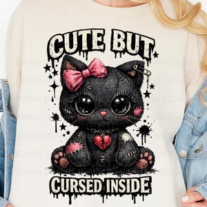 May include: Cream-colored sweatshirt featuring a black cat graphic with a pink bow, heart, and patches. The text "CUTE BUT CURSED INSIDE" is printed above and below the cat illustration. The design includes stars and ink splatters.