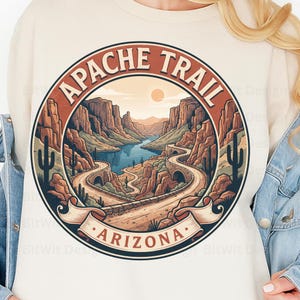 Op de afbeelding: Een crèmekleurig sweatshirt met een cirkelvormige afbeelding van de Apache Trail in Arizona. Het ontwerp toont een kronkelende weg door een woestijnkloof met een rivier, cactussen en een zon, omlijst door de tekst Apache Trail en Arizona.