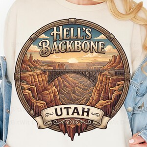 Peut inclure: Un sweat-shirt couleur crème avec une impression graphique circulaire représentant un paysage de canyon et un pont. Le texte indique HELL'S BACKBONE en haut et UTAH sur une bannière en bas. L'illustration inclut des falaises rocheuses.