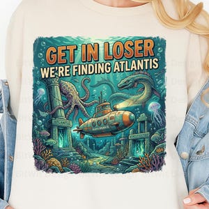Könnte beinhalten: Ein cremefarbenes Sweatshirt mit einem Grafikdruck einer Unterwasserszene mit U-Boot, Krake, Seeschlange und antiken Ruinen. Der Text "GET IN LOSER WE'RE FINDING ATLANTIS" ist in orangefarbenen und weißen Buchstaben über der Illustration zu sehen.
