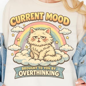 May include: A cream-coloured sweatshirt with a cartoon cat on a cloud, set against a rainbow. The text "CURRENT MOOD" arches above the cat, with "BROUGHT TO YOU BY OVERTHINKING" below. The cat has a disgruntled expression.