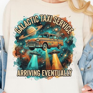 May include: A cream-colored t-shirt featuring a retro-style illustration. The design includes a vintage orange taxi cab floating in space, surrounded by planets, stars, and UFOs. Text reads "GALACTIC TAXI SERVICE ARRIVING EVENTUALLY."