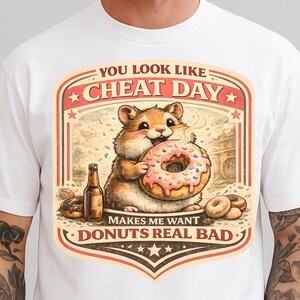 May include: White t-shirt featuring a cartoon hamster holding a donut with pink frosting and sprinkles. The graphic includes a beer bottle and the text "YOU LOOK LIKE CHEAT DAY MAKES ME WANT DONUTS REAL BAD."
