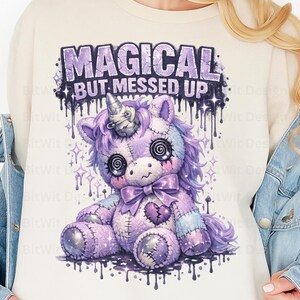 May include: A cream-colored sweatshirt featuring a purple unicorn design with the text "MAGICAL BUT MESSED UP." The unicorn has spiral eyes, a bow, and a stitched heart. The design includes dripping paint and star accents.