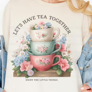 May include: Cream-colored sweatshirt featuring a stack of three teacups in pastel colors, adorned with floral arrangements. The text "LET'S HAVE TEA TOGETHER" arches above the cups, with "ENJOY THE LITTLE THINGS" below.