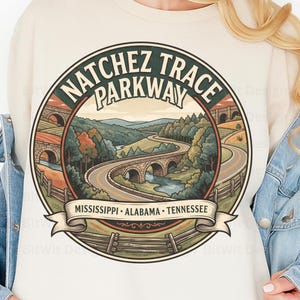 May include: A cream-colored sweatshirt featuring a circular graphic of the Natchez Trace Parkway. The illustration depicts a winding road, stone bridges, and a river through a forest landscape. The text reads Natchez Trace Parkway and Mississippi, Alabama, Tennessee.