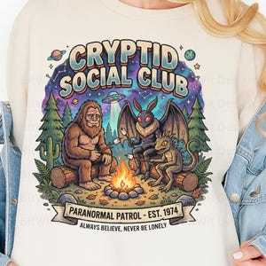 May include: Cream-colored sweatshirt featuring a colorful graphic of cryptids around a campfire. The design includes a Bigfoot, a Mothman, and a reptilian creature roasting marshmallows. Text reads "CRYPTID SOCIAL CLUB" and "PARANORMAL PATROL - EST. 1974."
