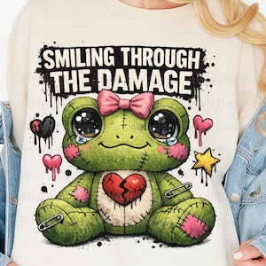 Könnte beinhalten: Ein grüner Plüschfrosch mit rosa Schleife, gebrochenem Herzen und dem Text "SMILING THROUGH THE DAMAGE". Der Frosch hat große Augen mit Tränen, aufgenähte Flicken und ist mit Herzen und einem Stern verziert.