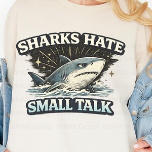 May include: Cream-colored sweatshirt with a graphic of a shark in blue and gray, emerging from water. The text "SHARKS HATE SMALL TALK" is above and below the shark illustration in black and teal.